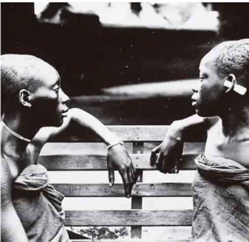 Fotografía en blanco y negro de dos niños negros sentados en una banca de madera, mirándose, en un entorno precario.