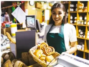 Vendedora en un negocio con pan en canasta y caja registradora, ilustrando las actividades económicas.