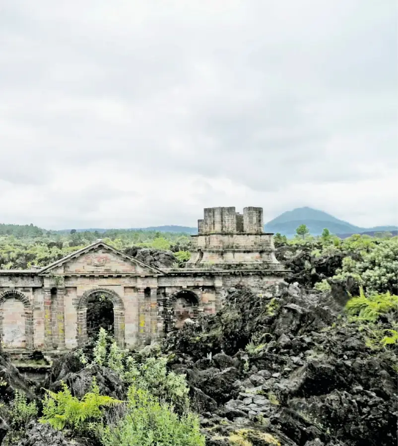 Fotografía de las ruinas de un templo de cantera con un frontón y una torre, rodeadas de rocas volcánicas negras y vegetación verde, con un cerro al fondo bajo cielo nublado.