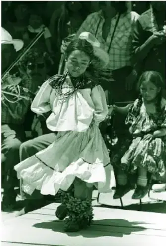Fotografía en blanco y negro de una niña guarijio bailando, con falda y blusa de manta bordada, collares de cuentas, cascabeles o coyoles en los tobillos y huaraches; al fondo otra niña sentada con vestido más oscuro y personas que rodean la danza.