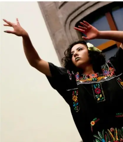 Fotografía de una bailarina con los brazos extendidos en un movimiento expresivo, vistiendo un traje negro bordado con motivos coloridos, ilustrando los códigos de la danza.