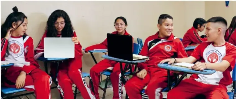 Fotografía de estudiantes de secundaria con uniforme rojo, sentados en su aula con laptops y cuadernos, trabajando colaborativamente en la preparación de su video proyecto.