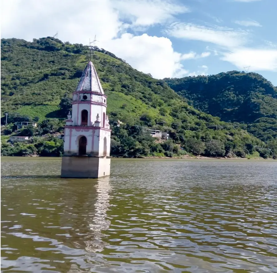 Fotografía de la torre blanca y rosada de la iglesia de la Asunción de María saliendo del agua de la presa, rodeada de cerros verdes cubiertos de vegetación y cielo con nubes.