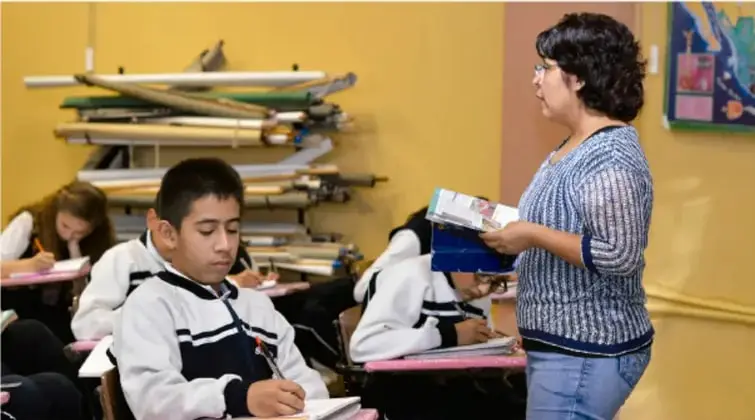 Fotografía de un aula: estudiantes con uniforme escriben en sus cuadernos mientras una docente de pelo corto oscuro, con gafas y suéter a rayas, sostiene un libro y supervisa la actividad; al fondo hay material deportivo y un mural.