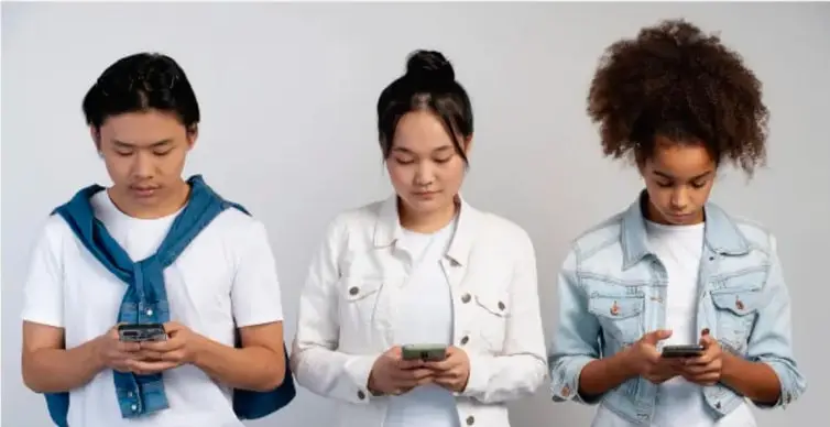 Fotografía de tres adolescentes (dos mujeres y un hombre) de pie y mirando sus teléfonos celulares, representando el uso cotidiano de las redes sociales.