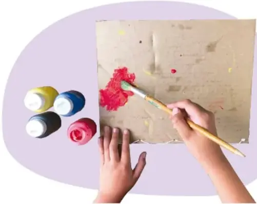Manos pintando un cartón con pincel; junto hay frascos de pintura de colores.