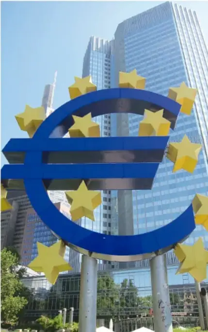 Escultura monumental del símbolo del euro (€) de color azul con estrellas amarillas, ubicada frente a un edificio moderno de oficinas, probablemente en Fráncfort, sede del Banco Central Europeo.