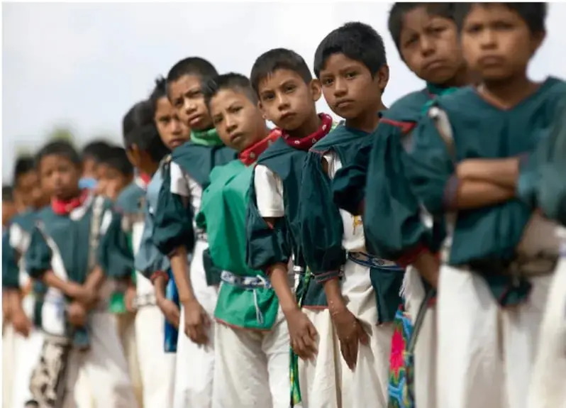 Fila de niños y niñas indígenas de pie, con camisas blancas, prendas verdes y azules, fajas tejidas y pañuelos rojos; al fondo un paisaje abierto.