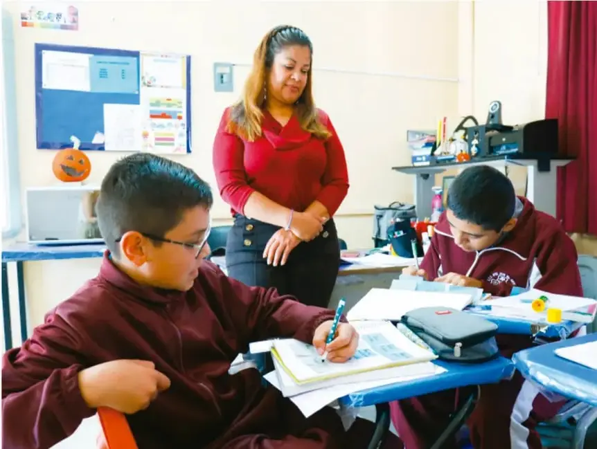 Fotografía de un aula: una maestra de pie observa a dos estudiantes uniformados que escriben en sus cuadernos sobre los escritorios.