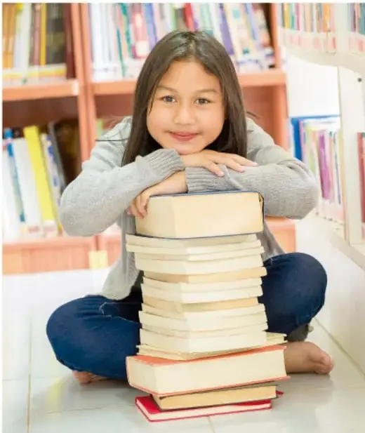 Niña sonriente sentada en el suelo con una torre de libros sobre las piernas, en una biblioteca.