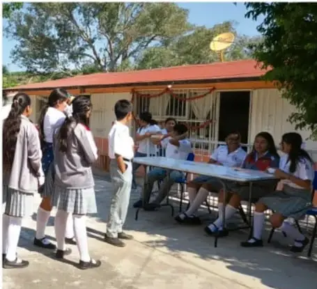 Fotografía de estudiantes de telesecundaria realizando una asamblea escolar al aire libre, algunos sentados en mesas y otros de pie participando.
