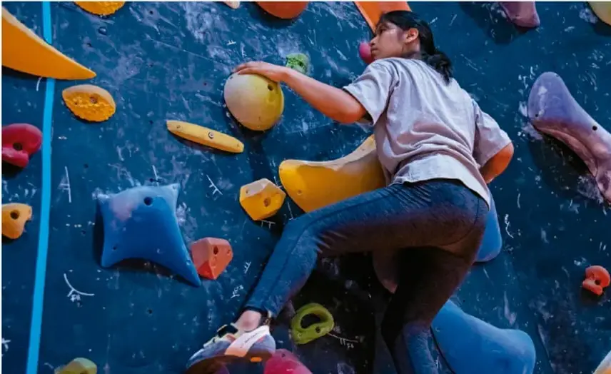 Fotografía de una estudiante practicando escalada en un muro de escalada deportiva con presas de colores.