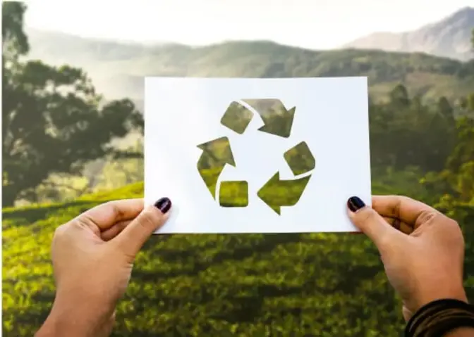 Fotografía de dos manos sosteniendo una hoja de papel con el símbolo de reciclaje recortado, sobre un fondo natural de bosque y montañas.