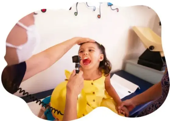 Fotografía de una niña con vestido amarillo abriendo la boca mientras un médico revisa su garganta con otoscopio y la otra mano le palpa la frente; consulta médica pediátrica.