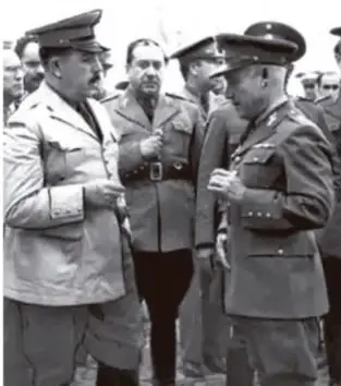 Fotografía en blanco y negro de tres militares conversando; uno de ellos viste uniforme claro y los otros dos uniformes oscuros con gorra.