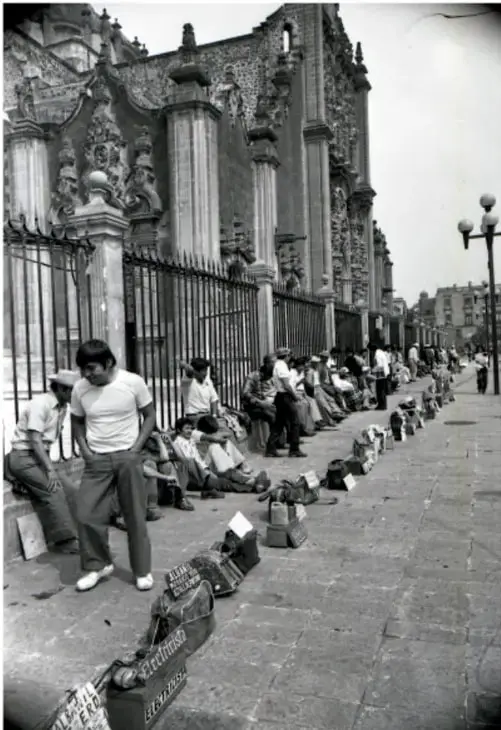 Fotografía en blanco y negro de una calle con varias personas sentadas en el suelo junto a una pared, aparentemente vendiendo productos. Al fondo se observa un edificio con arquitectura colonial y una reja.