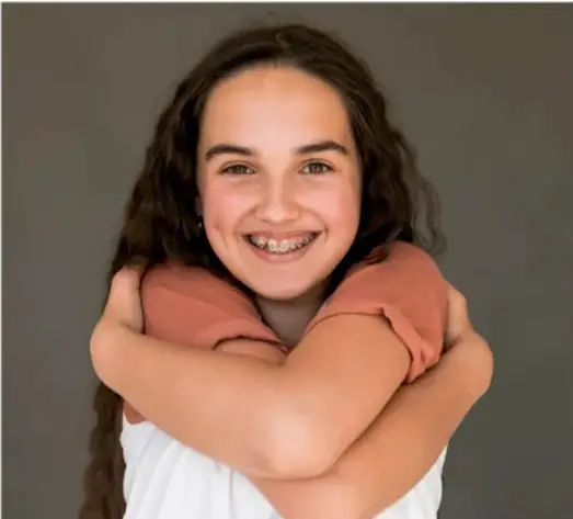 Fotografía de una joven sonriente con el cabello largo, abrazándose a sí misma con los brazos cruzados sobre el pecho, sobre un fondo neutro; representa la autoaceptación y el bienestar personal.
