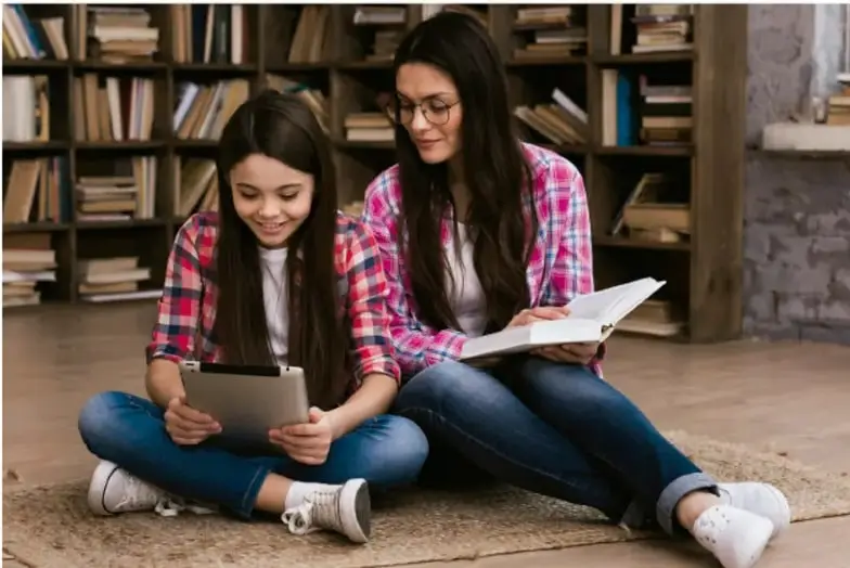 Fotografía de dos jóvenes sentadas en el piso de una biblioteca, una usa una tableta digital y la otra lee un libro, ilustrando el uso combinado de tecnología y fuentes tradicionales de información.