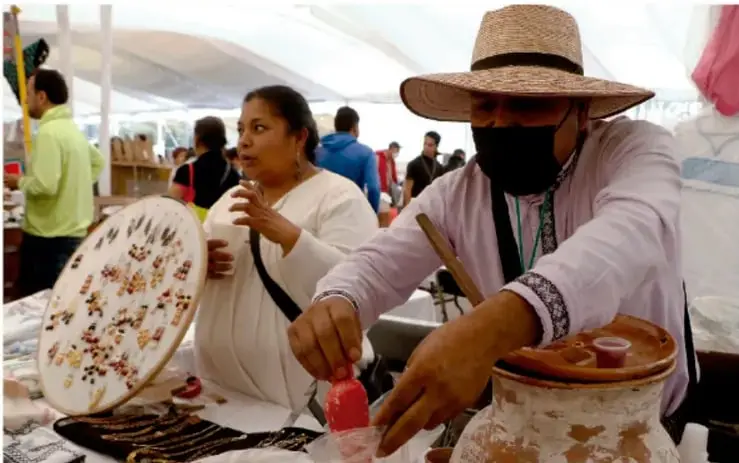 Fotografía de una muestra gastronómica: una mujer con blusa blanca sostiene un plato decorado con flores comestibles, mientras un hombre con sombrero de palma y cubrebocas sirve una bebida de una olla de barro.