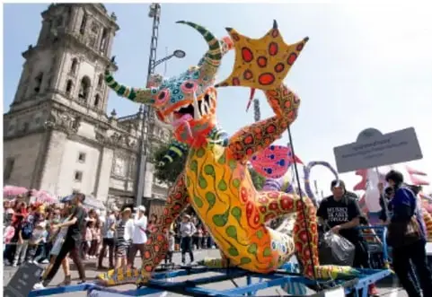 Registro fotográfico de una intervención artística en un espacio público: una gran figura de dragón de colores (papel maché) sobre una estructura, rodeada de personas y con un edificio histórico al fondo.
