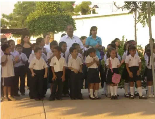 Grupo de estudiantes de primaria formados en el patio de la escuela, acompañados por una maestra y un adulto mayor.