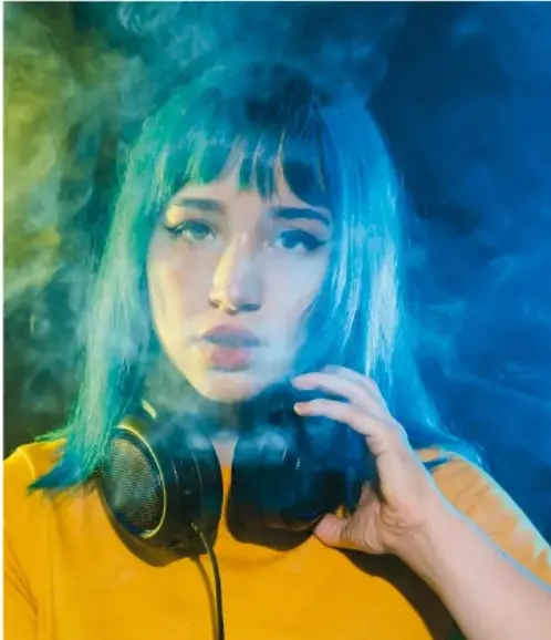 Foto de una joven con audífonos rodeada de humo de colores, representando el consumo de tabaco.