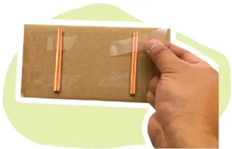 Mano pegando dos segmentos de popote sobre el cartón con cinta adhesiva.