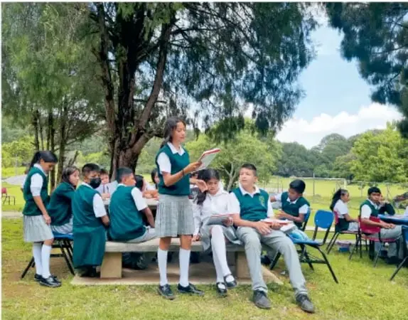 Grupo de estudiantes de telesecundaria con uniforme escolar conversando y trabajando al aire libre en un parque con árboles.
