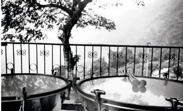 Fotografía en blanco y negro de tambores de percusión sobre un atril, con una reja y un árbol al fondo, en un entorno de sierra.