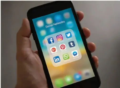 Fotografía de una mano sosteniendo un teléfono inteligente que muestra una carpeta llamada 'Social Networks' con íconos de Facebook, Instagram, Twitter, Pinterest, Tumblr, LinkedIn y WhatsApp.