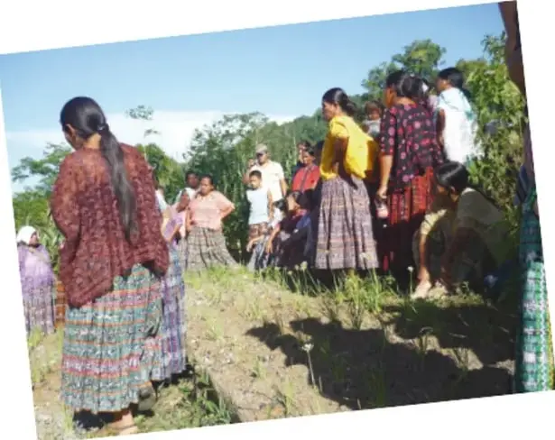 Fotografía de una asamblea comunitaria al aire libre: mujeres y hombres de pie y sentados en un terreno con vegetación, algunas personas con vestimenta tradicional.