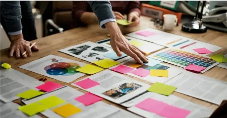 Fotografía de un equipo de trabajo colaborando alrededor de una mesa con notas adhesivas de colores, tarjetas, paletas de color y documentos de planeación de proyecto.
