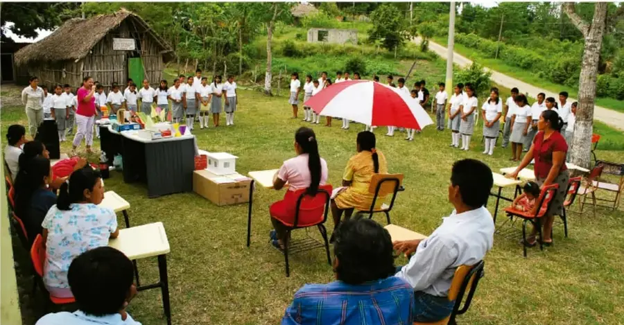 Fotografía de Jesús Eduardo Arteaga Flores (Tamaulipas): actividad escolar comunitaria al aire libre con estudiantes en formación, docentes y público sentado en sillas, carpas y materiales de trabajo sobre un pavimento, rodeados de árboles y una construcción de palma.