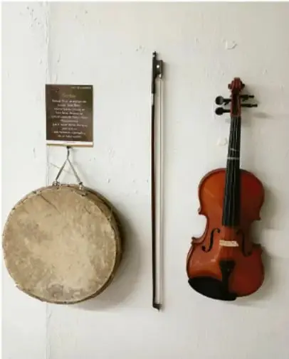 Fotografía de instrumentos musicales tradicionales colgados de una pared blanca: un tambor de piel con marco de madera, un arco para instrumento de cuerda y un violín, junto a una placa conmemorativa. Ejemplifica objetos regionales/artesanales que forman parte de un acervo artístico.