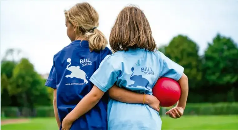 Fotografía de dos niñas de espaldas, con camisetas azules, tomadas por la cintura; una de ellas sostiene una pelota roja bajo el brazo.