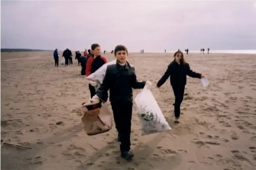 Grupo de estudiantes y adultos recogiendo basura en una playa, con bolsas en las manos.