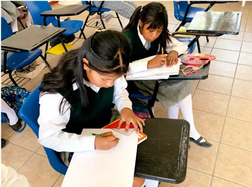 Fotografía de dos estudiantes de Telesecundaria con uniforme (chamarra verde y blanco) trabajando en sus pupitres: una de ellas dibuja o recorta sobre una hoja con tijeras y una forma geométrica color naranja, la otra escribe en su cuaderno; hay material didáctico (figuras geométricas de colores) sobre el pupitre, en un aula con pizarrón y pupitres azules.