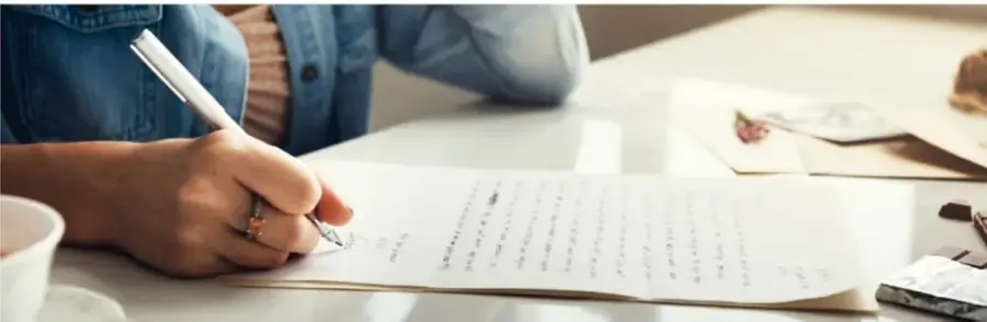 Fotografía de una persona escribiendo una carta a mano sobre una mesa, con taza y platos.