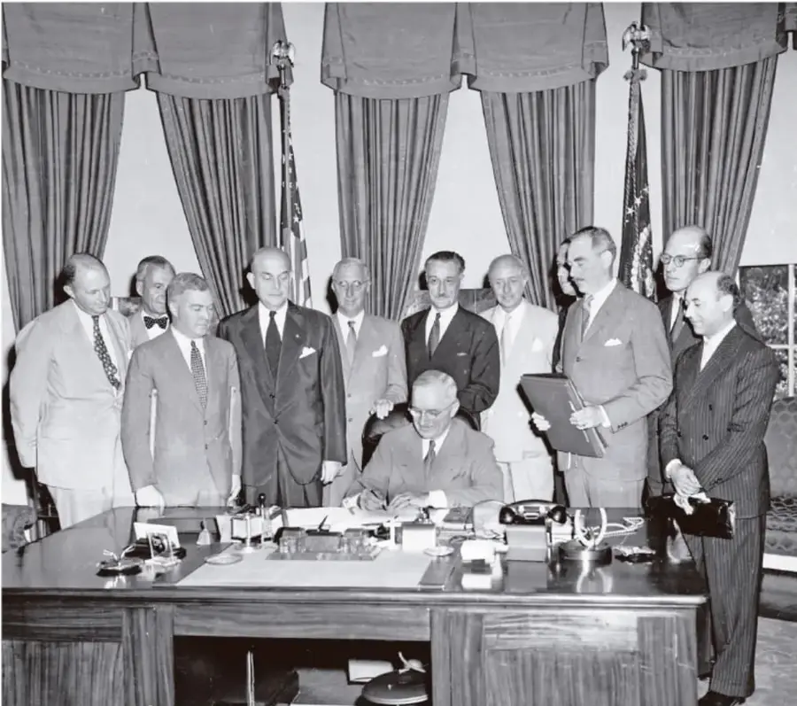 Fotografía en blanco y negro de un grupo de hombres en traje firmando un documento en una mesa, con cortinas al fondo. Parece ser la firma del Pacto de Varsovia.