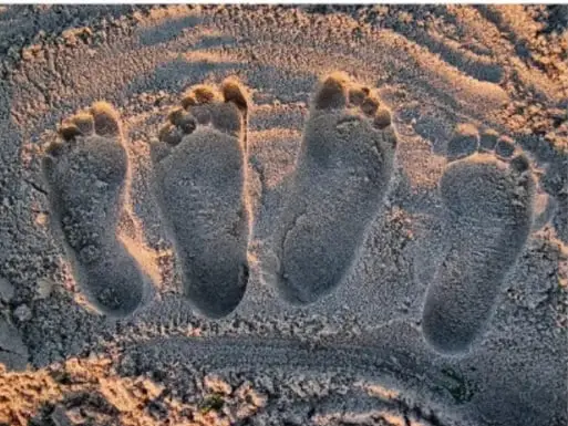 Fotografía de cuatro huellas de pies de bebé impresas en arena iluminada por la luz cálida del atardecer, en alusión a las huellas como identidad.