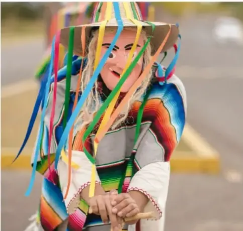 Fotografía de una persona con máscara pintada y sombrero decorado con listones de colores (huehuenche o danzante de carnaval), vestida con manta y sarape multicolor.