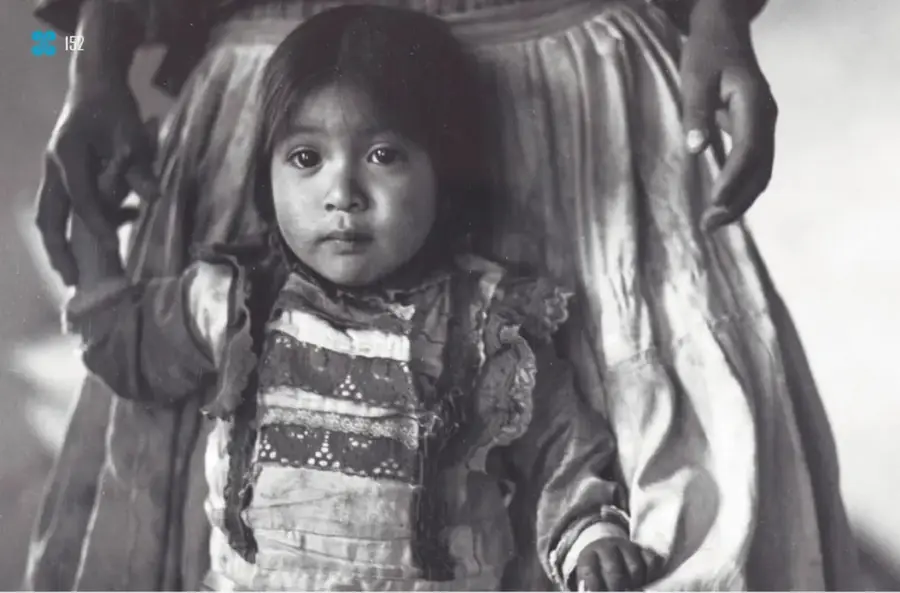 Fotografía en blanco y negro de una niña con vestido tradicional mirando a la cámara, junto a una mujer con mandil; El mandil, 1988, San Simón de la Laguna, Estado de México, de Mariana Yampolsky (1925-2002).