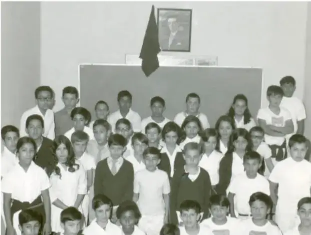 Fotografía en blanco y negro de un grupo numeroso de estudiantes de primaria, con uniforme escolar, posando frente a un pizarrón; al fondo se observa un retrato y una bandera.