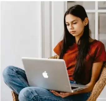 Fotografía de una estudiante adolescente sentada con una computadora portátil sobre las piernas, investigando o estudiando.