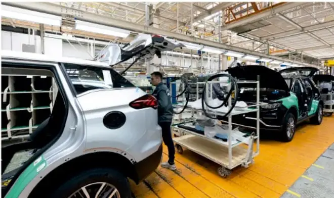 Fotografía de una planta armadora de automóviles: un trabajador junto a una carrocería blanca en una línea de ensamblaje con estructura metálica y suelo amarillo.