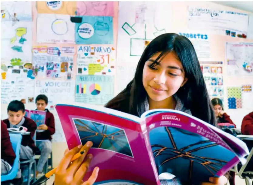 Estudiante leyendo un libro de la colección Ximhai en un aula con murales, carteles (Año Nuevo, Historia, El Voceo de cocina) y compañeros de fondo.