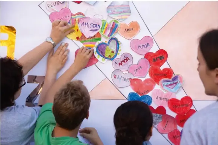 Manos de estudiantes pegando corazones de cartulina de colores con las palabras 'AMOR', 'AMISTAD' y 'RESPETO' sobre un mural en forma de cruz o escalera.