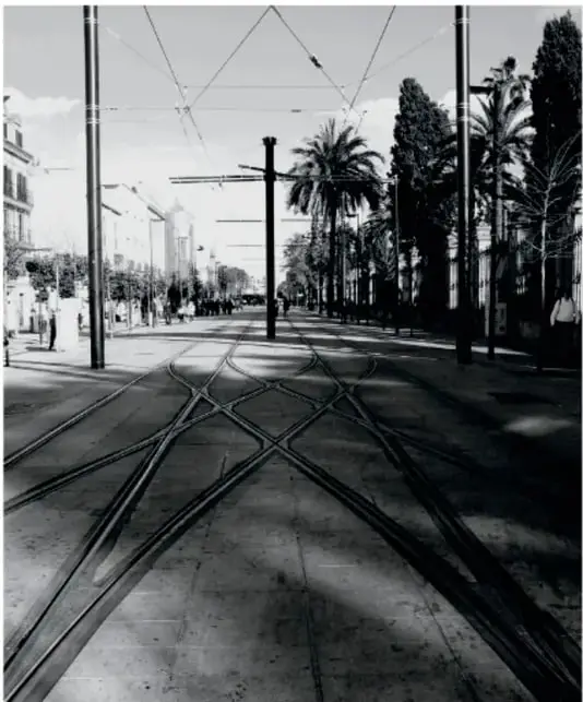 Fotografía en blanco y negro de una calle con vías de tranvía que se cruzan y se dirigen hacia un punto de fuga, con postes, árboles y palmeras a los lados.
