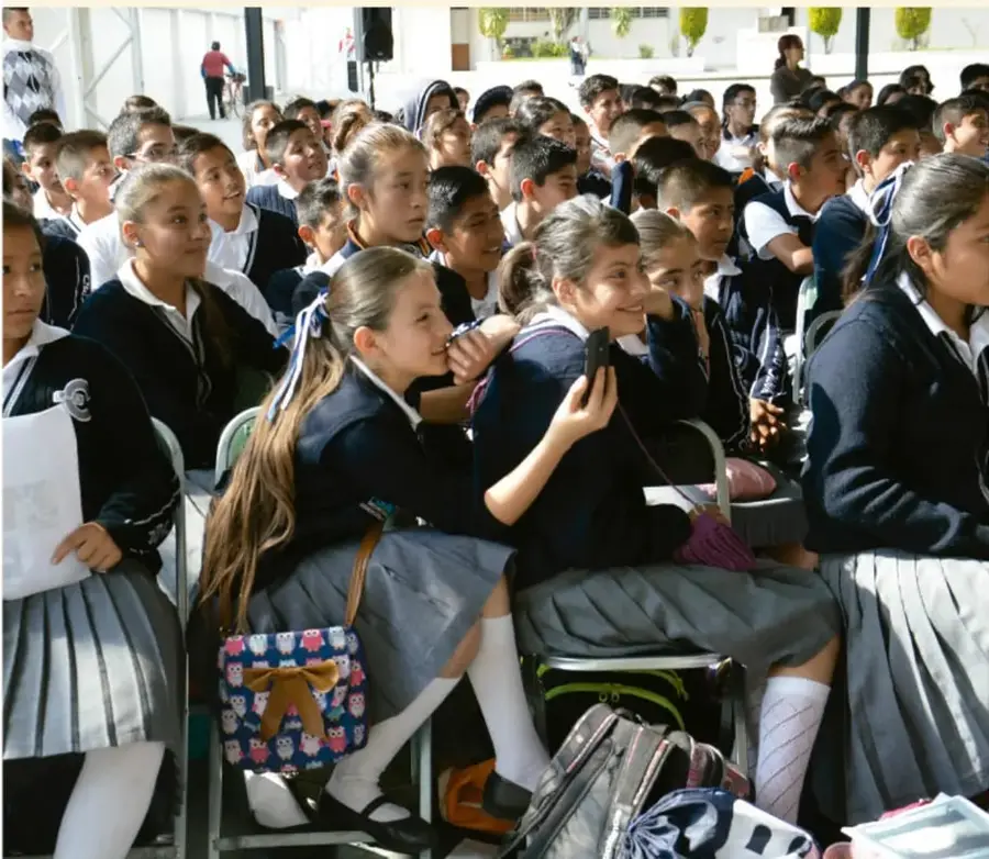 Fotografía de un grupo de estudiantes de secundaria con uniforme (chamarra azul y falda gris plisada) sentados en sillas durante un acto escolar, varios conversan entre ellos y observan el evento.