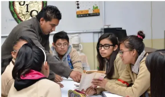 Fotografía de un grupo de estudiantes de telesecundaria y un docente reunidos alrededor de un mesa, leyendo y compartiendo un libro de texto en el aula.